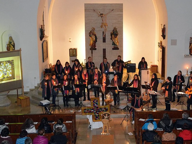 De Lumina - bei Ihrem Konzert in Mörnsheim am 26.11.2017 Chor mit bunten Schals um den Hals spielen auf Instrumenten und singen, mit brennenden Kerzen vor ihnen in einer Kirche mit weißen Wänden, mit dem Jesuskreuz und weiteren Gebilden von Heiligen im Hintergrund. Publikum auf dunkelbraunen Holzbänken sitzend.