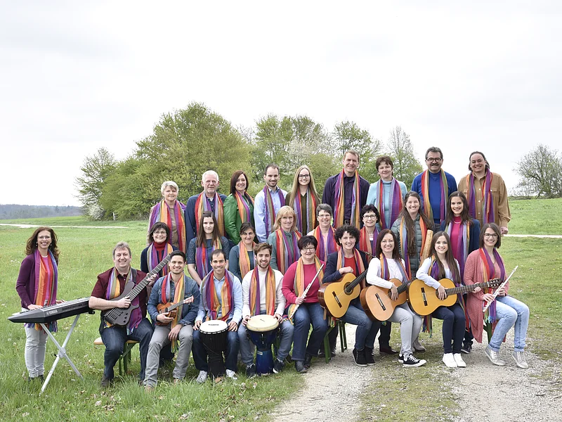 De Lumina - der Chor Gruppenfoto von 28 Personen mit Musikinstrumenten und bunten Schals um den Hals auf einer Wiese vor Bäumen bei bewölktem Himmel.