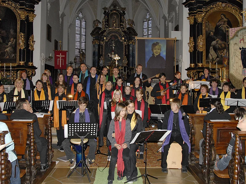 De Lumina - Im Jahr 2009 Chor mit bunten Schals singt und musiziert in einer Kirche vor Publikum auf braunen Holzbankreihen.