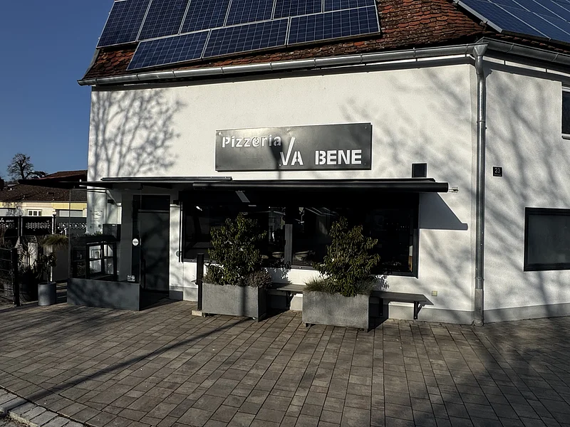 Pizzeria Va Bene - Bei schönem Wetter Essensmöglichkeit draußen vor der Pizzeria Außenansicht einer Pizzeria mit schwarzem Schild „Pizzeria VA BENE“, eine Eingangsbereich aus Glas, verdunkeltem großen Fenster mit Rollläden und zwei großen Pflanzkübeln davor. Vor der Pizzeria ein gepflasterter Gehweg. Auf dem Dach des Gebäudes viele Solarpanele.