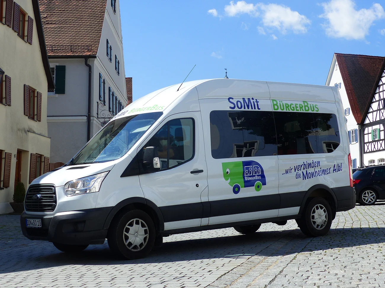 Weißer Kleinbus steht auf einem gepflasterten Platz, in der Monheimer Innenstadt . Auf dem Bus ist die Aufschrift "SoMit BürgerBus" und "wir verbinden die Monheimer Alb".