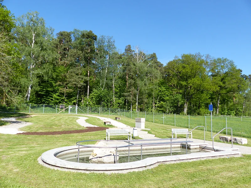 Rundes Kneippbecken in Monheim mit einem Geländer und einem Barfußpfad auf einer Wiese, umgeben von einer großen Grünfläche und Bäumen und Sitzmöglichkeiten. Ein Zaun im Hintergrund.