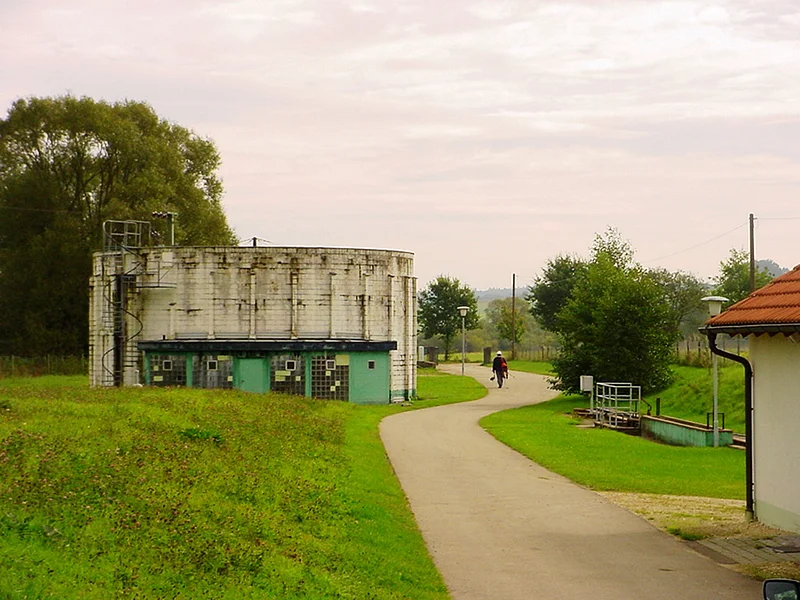 Die "alte" Kläranlage der Stadt Monheim von 1967 bis 2002 Zwei Gebäude links und rechts entlang eines geteerten Weges umgeben von grünen Wiesen und Bäumen. Eine Person mit Helm geht im Hintergrund auf dem Weg, weg vom Betrachter.