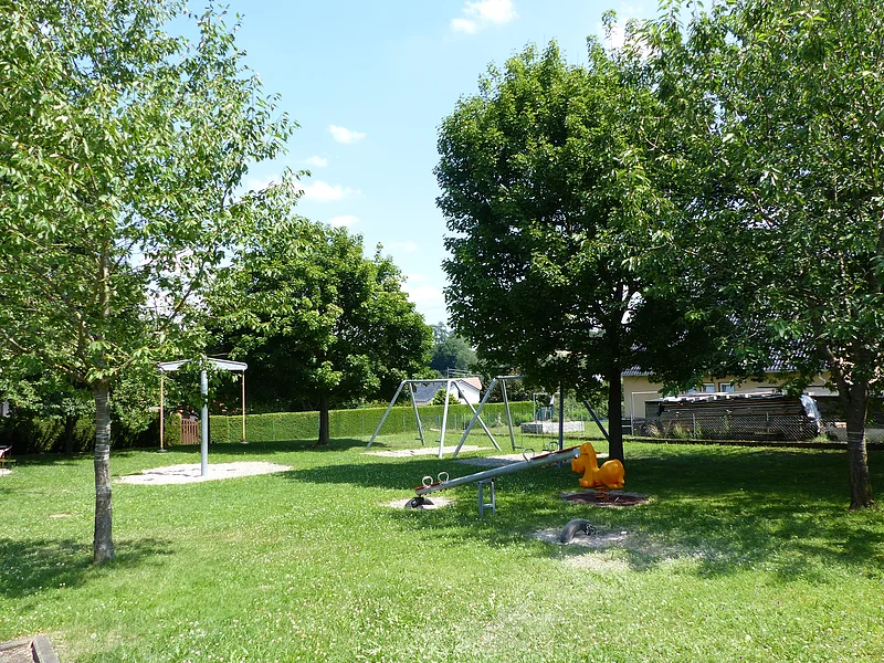 Spielplatz mit Wippen, Schaukeln und einem Sandkasten umgeben von grünen Bäumen auf einer Wiese bei Sonnenschein.