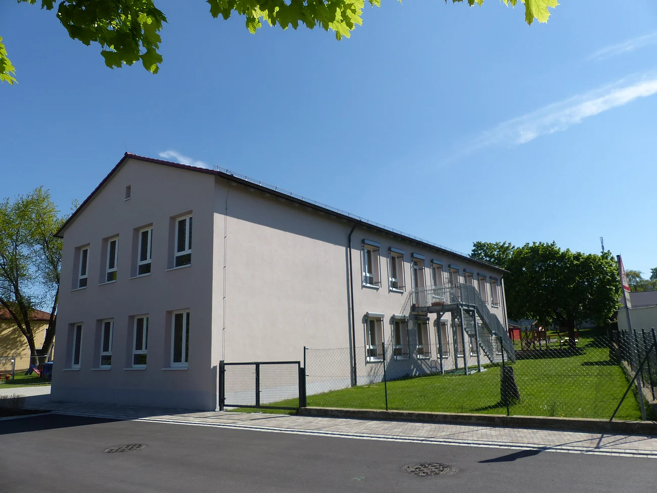 Städtischer Kindergarten in Monheim: ein zweistöckiges Gebäude mit weißer Fassade, Fenstern und Außentreppe, umgeben von einem Zaun und Bäumen. Rechts um das Gebäude ist ein Garten zu sehen. Vorderhalb des Gebäudes verläuft eine Straße.