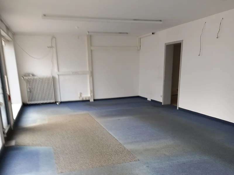 Leerer Raum mit blaugrauem Teppichboden und weißen Wänden, Heizkörper links hinten an der Wand und offener Tür rechts sichtbar.