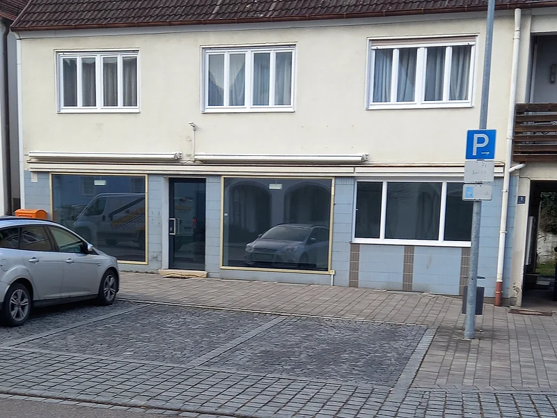 Zweistöckiges zweifarbiges Gebäude mit drei Fenstern oben und drei großen Schaufenstern unten. Ein parkendes Auto auf einem Parkplatz davor.