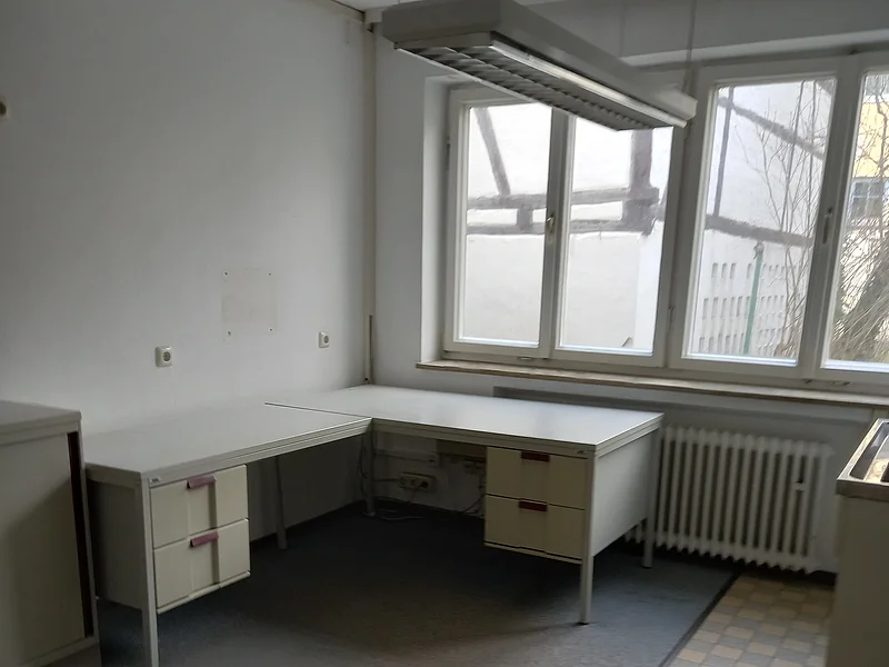 Leerer Büroraum mit Eckschreibtisch, Fenster, Heizkörper dahinter und hängender Deckenleuchte, sowie grauer Teppichboden und Fliesenbereich.