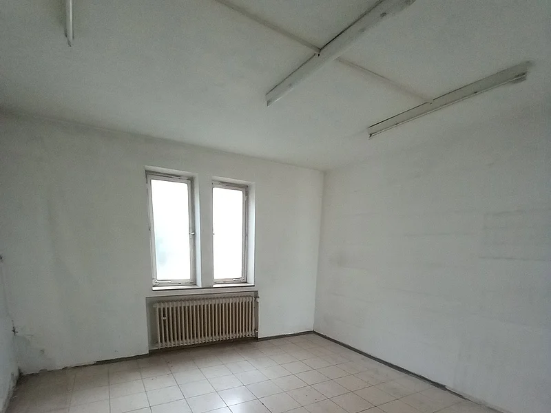 Leerer Raum mit weißen Wänden, zwei Fenstern, einem Heizkörper darunter und gefliestem Boden. An der Decke drei Leuchtröhren.