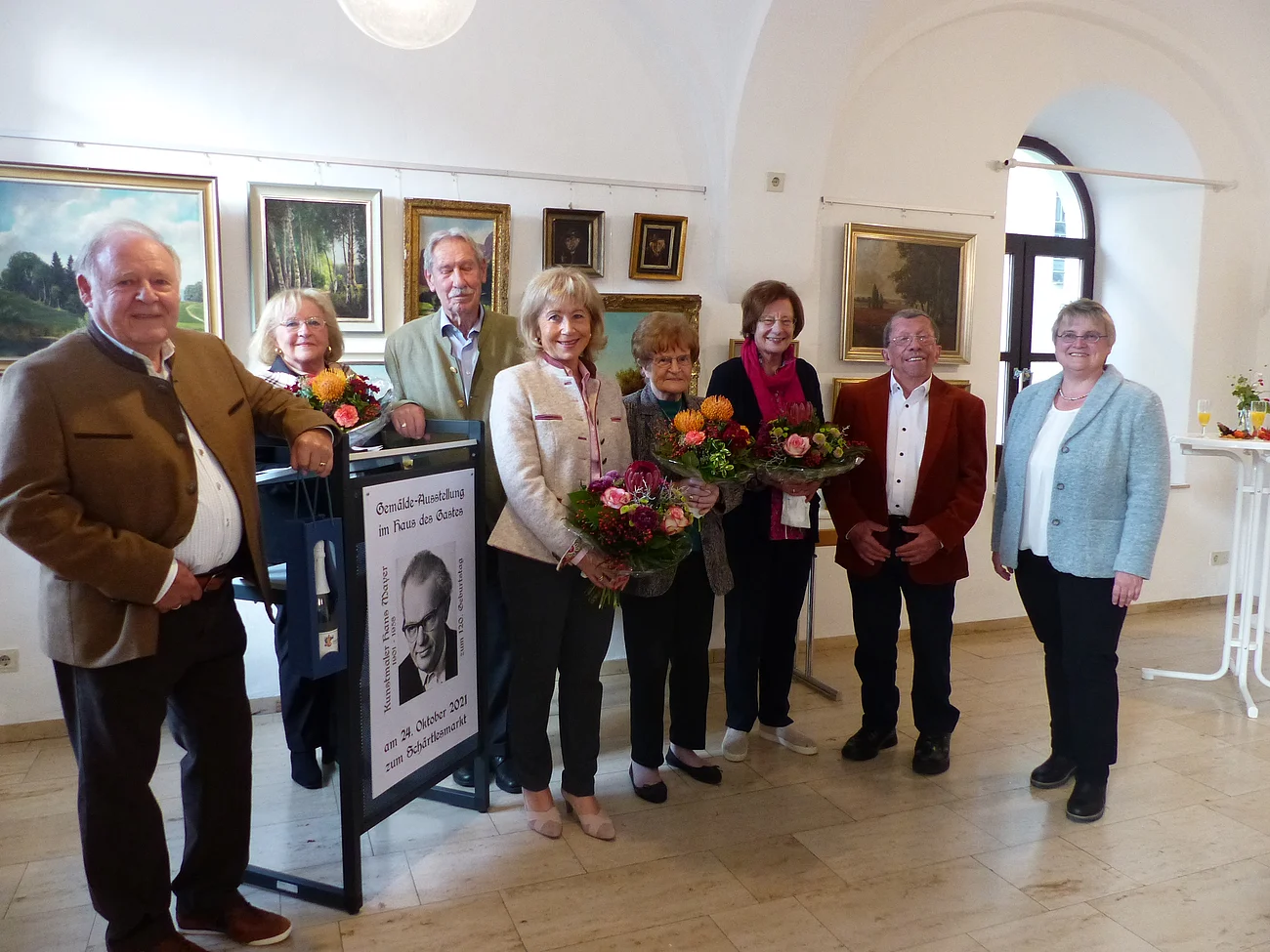Die Haupt-Akteure der Gemäldeausstellung zum 120. Geburtstag des Kunstmalers Hans Mayer Gruppe von acht Personen in einer Galerie, einige halten Blumensträuße. Im Hintergrund sind Gemälde an der Wand. Vor den Personen steht ein Aufsteller mit einem Portrait des Kunstmalers Hans Mayer aus Monheim.