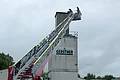 Feuerwehrwagen mit ausgefahrener Leiter vor einem Gebäude mit "Gerstner"-Schild. Auf der Leiter sind zwei Personen zu sehen, die Störche beringen. Im Hintergrund bewölkter Himmel.