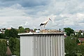 Ein Storch steht auf einem Nest auf einem hohen, metallischen Gebäude. Im Hintergrund sind Bäume, blauer Himmel und Häuser zu sehen.