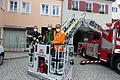 Drei Personen mit Schutzmasken stehen auf einer Feuerwehr-Hubarbeitsbühne vor einem roten Feuerwehrfahrzeug mit ausgefahrener Leiter. Im Hintergrund zwei farbige Gebäude und gepflasterter Platz vorne.