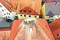Rotes Dach mit einem Storchennest und einem Storch darin, umgeben von bunten Häusern mit roten und grauen Dächern.