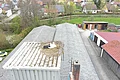 Weißstorch sitzt im Nest auf einem grauen Dach eines Sägemehlbunker in einer Kleinstadt, mit Häusern, Straßen, Bäumen und Wiesen im Hintergrund.