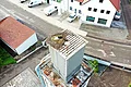 Zwei Störche in und an einem großen Nest auf einem hohen grauen Sägemehlbunker in einer Kleinstadt, umgeben von weiteren Häusern, mehreren Vans und einer Verkehrsstraße.