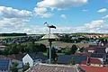 Ein Storch steht auf einem hohen Dach mit Blick auf Häuser und Felder einer Kleinstadt im Hintergrund.