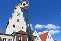 Feuerwehrleiterwagen mit ausgefahrener Leiter vor einem historischen Gebäude mit Torbogen und einem Turm dahinter. Auf dem Turm eine Sonnenuhr. Auf der Feuerwehrleiter stehen zwei Personen.