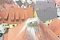 Dach mit einem Storchennest und einem Weißstorch, umgeben von weiteren Häuserdächern mit roten und dunklen Ziegeln in einem Ort.
