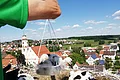 Eine Person wiegt einen jungen Storch in einer Schale über dem Nest auf einem Dach, mit Blick auf eine Stadtlandschaft und blauen Himmel. Im Nest liegen insgesamt drei Störche.