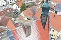 Weißstorch sitzt im Nest auf einem Dach mit roten Ziegeln in einem Dorf mit mehreren Häusern, bei bewölktem Himmel.