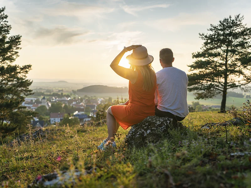 Ein Pärchen sitzt oberhalb von Monheim auf einem Stein umgeben von grüner Wiese und eingerahmt von zwei Bäumen. Die beiden genießen den Sonnenuntergang und den schönen Blick auf Monheim.