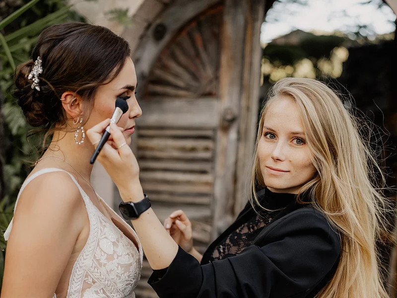 Laura Burlefinger - Make-up Artist & Hairstylist Monheim Frau trägt schwarzen Blazer und schminkt eine Braut in weißem Spitzenkleid vor Holztür draußen.