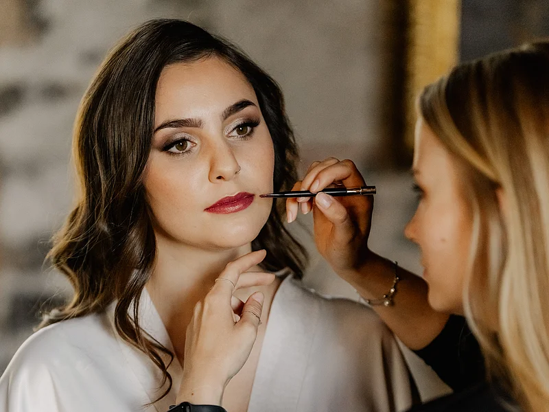 Laura Burlefinger - Make-up Artist & Hairstylist Monheim Frau trägt weißen Seidenbademantel, während eine andere Frau im schwarzem Blazer Lippenstift auf ihre Lippen aufträgt.
