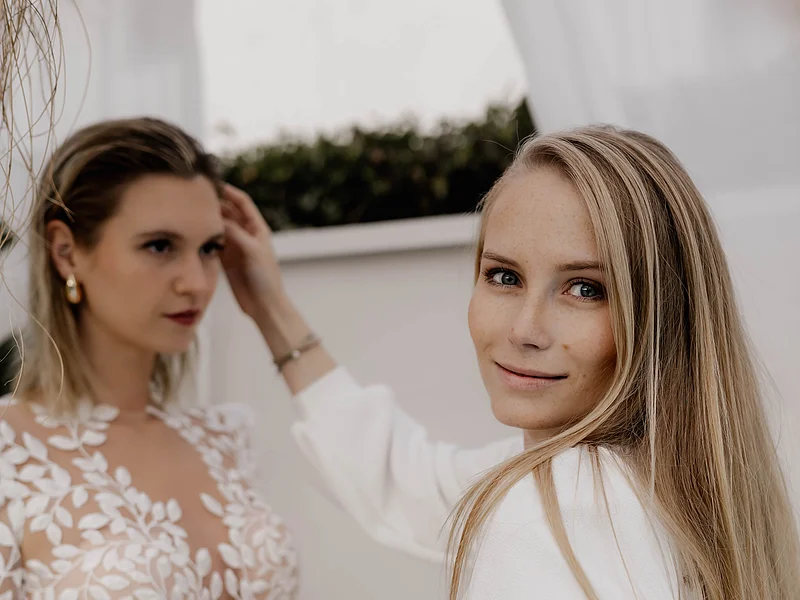 Laura Burlefinger - Make-up Artist & Hairstylist Monheim Zwei Frauen stehen nebeneinander, eine trägt ein weißes Hochzeitskleid mit Spitze, die andere hält einen Make-up-Pinsel und schaut in die Kamera.