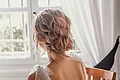 Laura Burlefinger - Make-up Artist & Hairstylist Monheim Frau mit hochgestecktem Haar und weißem, perlenbesetztem Kleid sitzt auf Holzstuhl vor Fenster mit weißen Vorhängen.