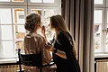 Laura Burlefinger - Make-up Artist & Hairstylist Monheim Frau trägt Seidenrobe sitzt auf Stuhl, während andere Frau ihr Make-up aufträgt vor großem Fenster.