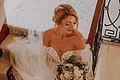 Laura Burlefinger - Make-up Artist & Hairstylist Monheim Braut in weißem Spitzenkleid mit Blumenstrauß steht auf Treppe und schaut nach unten.