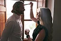 Laura Burlefinger - Make-up Artist & Hairstylist Monheim Frau trägt einem Mann mit Pinsel im Gesicht Make-up auf, beide vor offenem Fenster stehend.
