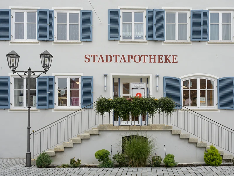 Gebäude mit blauen Fensterläden, rotem Schriftzug "STADTAPOTHEKE", einer Treppe und einer Laterne mit einem bepflanzten Beet davor.