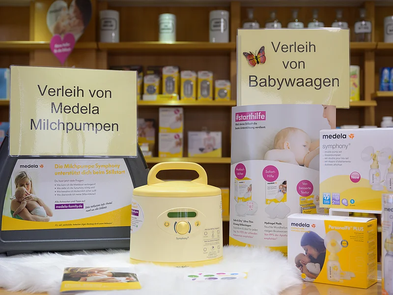 Präsentationstisch in einer Apotheke mit Einer gelben Milchpumpe der Firma "Medela", daneben ein Tragekoffer dazu und ein Schild mit dem Schriftzug "Verleih von Medela Milchpumpen". Ein weiteres Schild mit dem Schriftzug "Verleih von Babywaagen" rechts daneben, mit Produkten und Informationsmaterial auf einem Tisch.