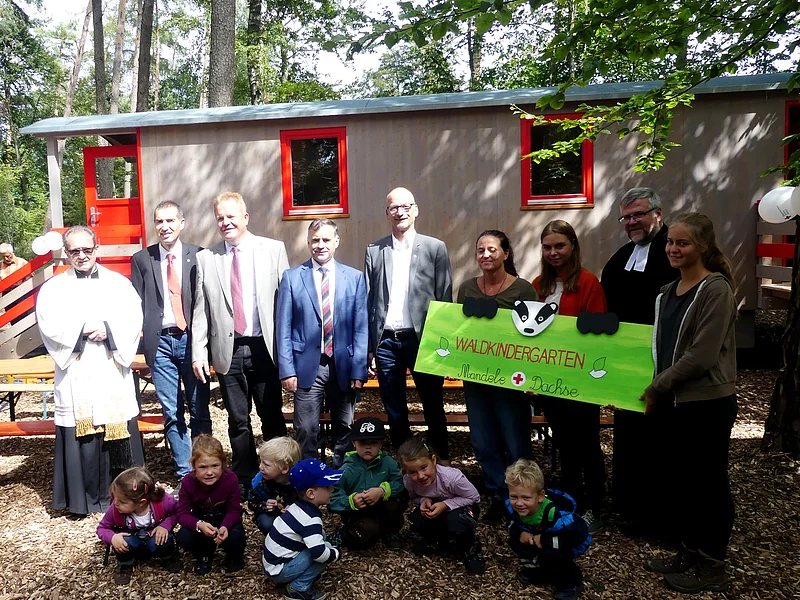 Waldkindergarten Mandele Dachse in Monheim eröffnet Eine Gruppe von Erwachsenen und Kindern steht bzw. kniet vor einem Bauwagen im Wald, drei Personen halten ein gestaltetes Schild mit der Aufschrift "Waldkindergarten Mandele Dachse" in den Händen.