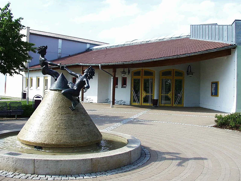 Stadt- und Mehrzweckhalle Monheim Kegelförmiger Steinbrunnen mit Wasser gefüllt, zwei spielende Kinderfiguren aus Metall oben drauf vor einem Gebäude mit gelben Türen und rotem Dach, auf einem gepflastertem Gebäude. Links im Bild ein weiteres angrenzendes hohes Gebäude bei blauem Himmel.