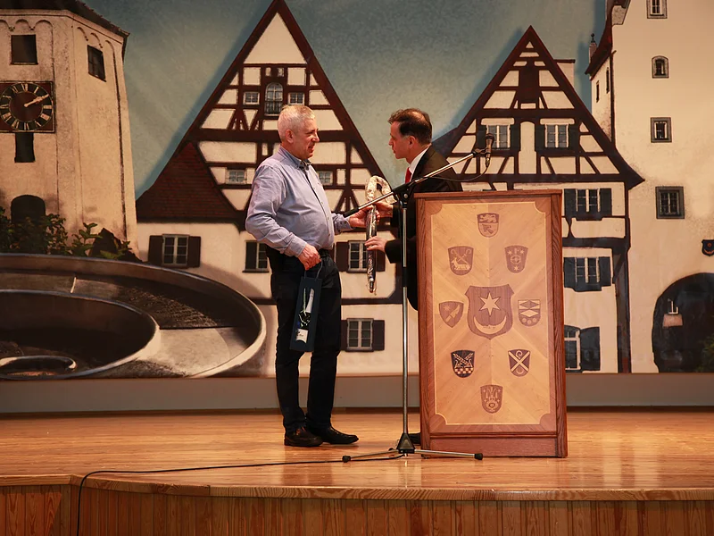 Jubiläumsveranstaltung "25 Jahre Stadthalle Monheim" - Hausmeister Josef Berkmüller wurde von Günther Pfefferer für seinen unermüdlichen Einsatz in der Stadthalle geehrt Zwei Männer auf Bühne mit einem Rednerpult, einer übergibt ein Geschenk, Im Hintergrund Wandbild mit Fachwerkhäusern und Uhrturm.