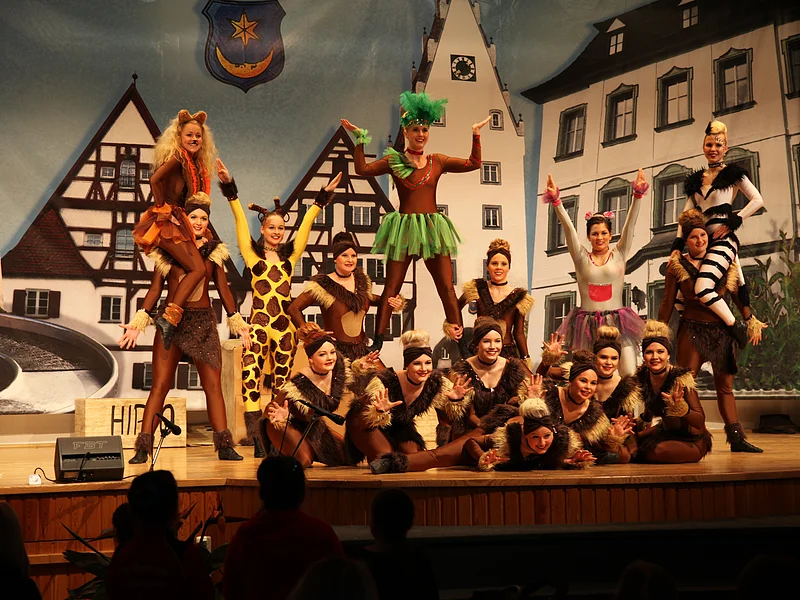 Jubiläumsveranstaltung "25 Jahre Stadthalle Monheim" - die FG Gailachia beeindruckte die Zuschauer mit den Tanzmäusen, der Mini-, Teenie- und Midigarde sowie der Prinzengarde um dem Zwillingstanzpaar Verkleidete Theatergruppe in Tierkostümen posiert auf einer Bühne vor Kulisse mit Fachwerkhäusern und einem Uhrturm.