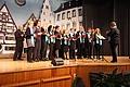 Jubiläumsveranstaltung "25 Jahre Stadthalle Monheim" - der Liederkranz Monheim mit den Chören Gemischter Chor, Jugendchor und Kinderchor unterhielt die Gäste mit Musik Chor mit Sängerinnen und Sängern auf einer Bühne, dirigiert von einer Person vorderhalb, alle mit blauen Schals und Notenheften. Im Hintergrund ein Aushang mit der Abbildung einer Stadt.