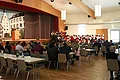 Jubiläumsveranstaltung "25 Jahre Stadthalle Monheim" - die Stadtkapelle und die Jugendkapelle Monheim sorgten für beste musikalische Unterhaltung Veranstaltung in einem Saal mit Publikum an Tischen, ein Redner steht am Podium auf der Bühne und einer Blaskapelle in rot-weißer Tracht rechts davon.