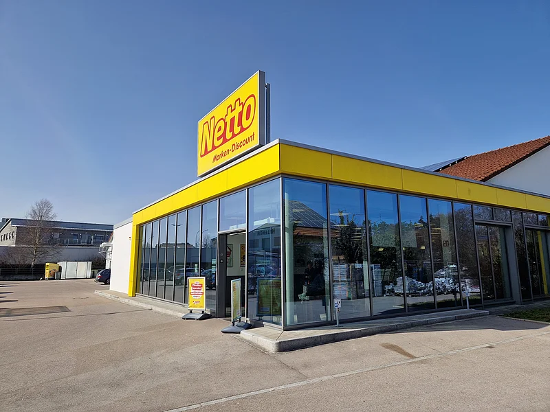Gebäude eines Netto Marken-Discounts mit einer Glasfassade, gelbem Fassadenband und dem Schriftzug "Netto" oben auf dem Gebäude in rot/gelb. Großer Parkplatz bei klarem Himmel.