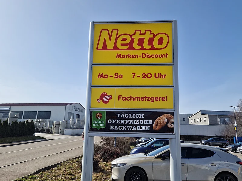 Schild mit Öffnungszeiten und Angeboten von Netto Marken-Discount an einem Parkplatz, an einer Straße, bei klarem Himmel.