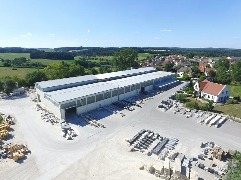 Marmorwerk Böswald in Weilheim Luftaufnahme eines großen Industriegebäudes mit Lagerplatz im Freien und angrenzenden Wohnhäusern in ländlicher Umgebung bei klarem Himmel.