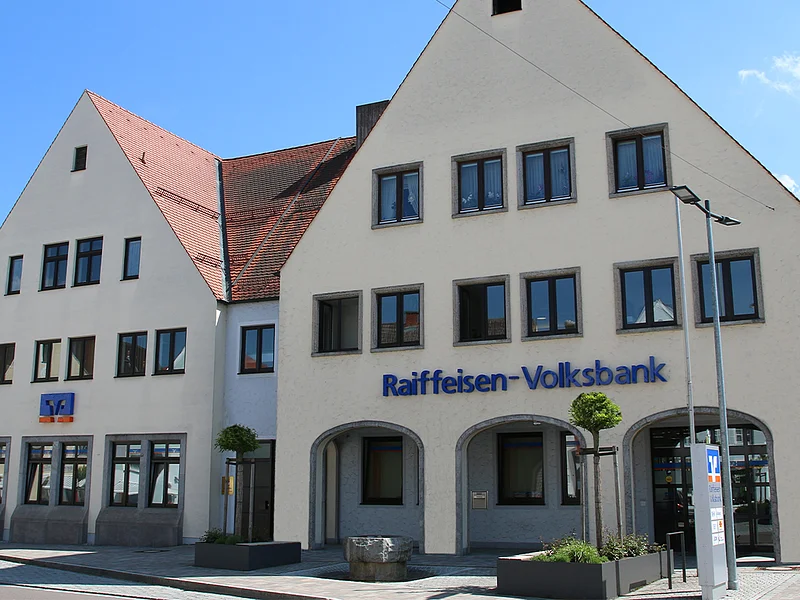 Zwei miteinander verbundene Gebäude mit Giebeldach und auf einem davon der Schriftzug „Raiffeisen-Volksbank“ bei klarem blauen Himmel, Straßenlaterne, einem Baum und  einem bepflanzten schwarzen Beet davor. Vor dem Gebäude verläuft eine Straße.
