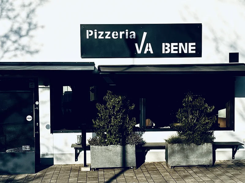 Pizzeria Va Bene - Außenansicht der Pizzeria Außenansicht einer Pizzeria mit schwarzem Schild „Pizzeria VA BENE“, eine Eingansbereich aus Glas, verdunkeltem großen Fenster mit Rollläden und zwei großen Pflanzkübeln davor. Vor der Pizzeria ein gepflasterter Gehweg.