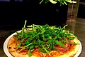 Pizzeria Va Bene - hier werden alle Gerichte frisch zubereitet Hand legt Rucola auf eine belegte Pizza mit Schinken auf weißem Teller in Küche.