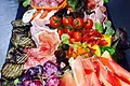 Pizzeria Va Bene - Antipasti vom Feinsten Antipasti-Platte auf schwarzer Unterlage mit Trauben, Tomaten, Melone, verschiedenen Wurst- und Käsesorten sowie gegrilltem Gemüse auf Salatblättern.