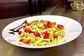 Pizzeria Va Bene - Pasta vom Feinsten Teller mit dampfender Pasta, Tomatenstücken, Pilzen und Kräutern, dekoriert mit Balsamico-Soße steht auf einem braunem Holztisch.