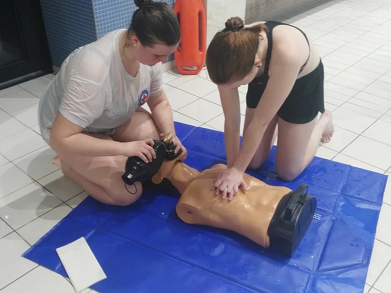 Zwei Frauen üben Herz-Lungen-Wiederbelebung an einer Übungspuppe auf einer blauen Matte in einem Schwimmbad.
